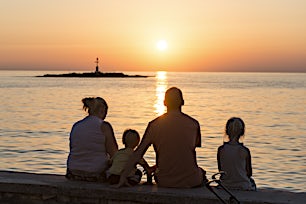 Aurinko laskee horisonttiin valaisten taivaan oranssiksi, perheen seuratessa tapahtumaa rannalta.