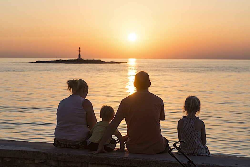 Aurinko laskee horisonttiin valaisten taivaan oranssiksi, perheen seuratessa tapahtumaa rannalta.