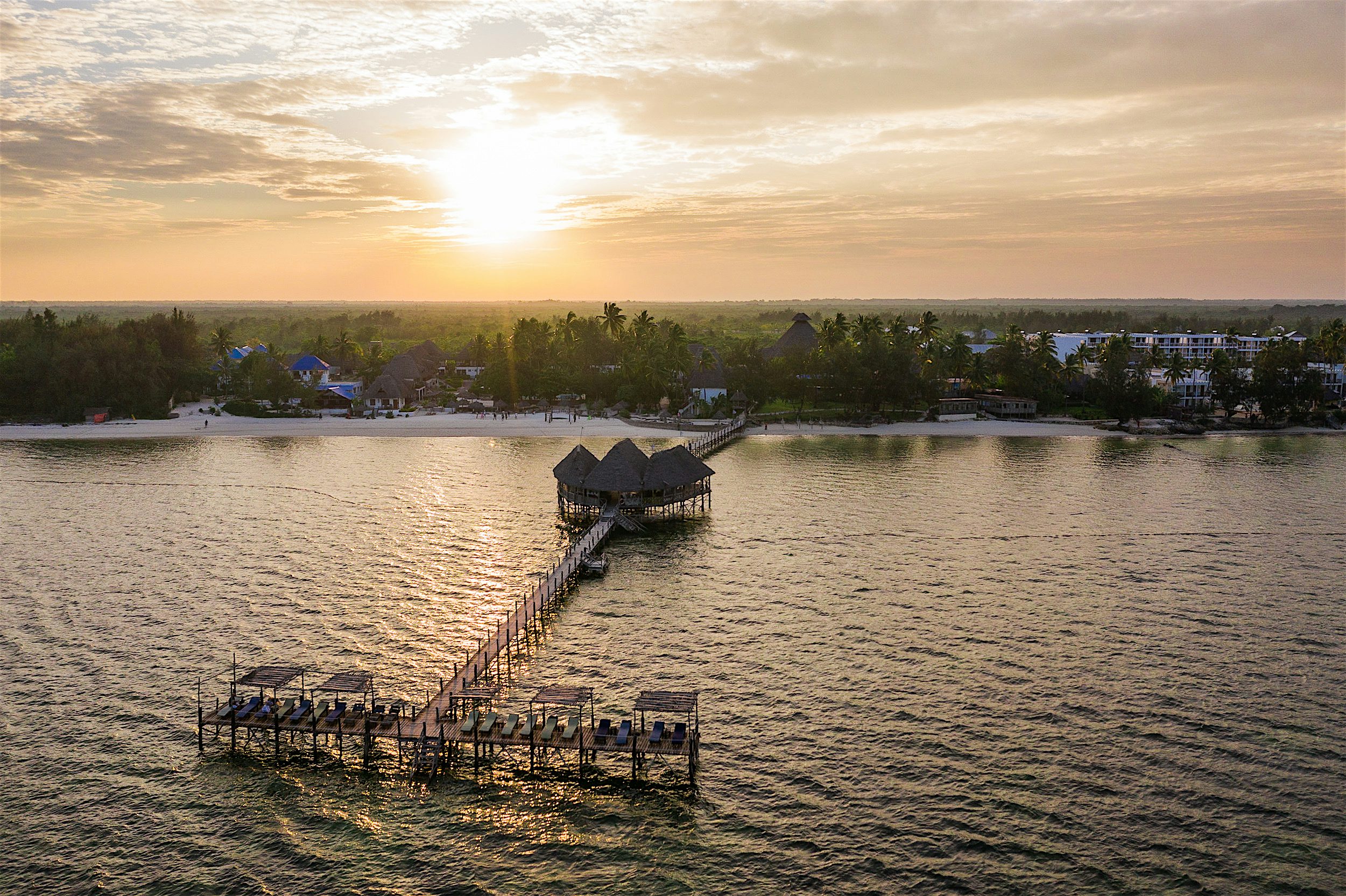 Pitkä laituri, jonka päässä aurinkotuoleja ja keskellä katettu osa. Ilta-aurinko heijastuu merestä ja taustalla on hiekkaranta ja matala hotellirakennus.